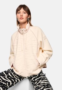 Cream zip-up jacket with textured stripes, high collar, and two front pockets. Paired with black and white zebra-striped pants.