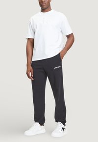 Weißes Baumwoll-T-Shirt mit geprägt Logo, kombiniert mit schwarzen Jogginghosen mit weißem Text. Modell trägt weiße Sneakers. Lässiges Outfit.