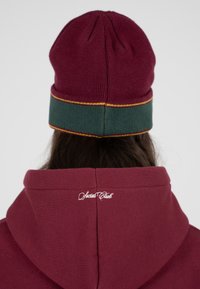 Bordowy dzianinowy beanie z zielonym paskiem i żółtą nitką. Ciemnobordowa bluza z kapturem z białym haftem "Social Club" u szyi.