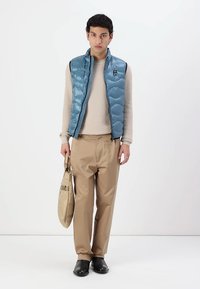 Blå polstret vest med glidelås, beige teksturert genser, beige bukser, og svarte sko. Modellen holder en beige skulderveske.