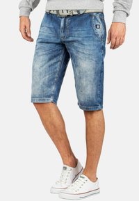Shorts en denim bleu avec un délavage, dotés d'une ceinture, de poches latérales et d'un patch logo à l'avant. Portés avec des baskets blanches.