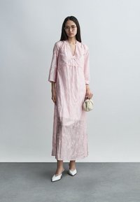 Γυναίκα που φοράει ένα ροζ μακρύ φόρεμα με κεντήματα, λευκές μυτερές γόβες, γυαλιά και κρατά μια μικρή μπεζ τσάντα χειρός μπροστά σε απλό φόντο.