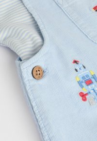 Blaue Cord-Overalls mit gesticktem Robotermotiv. Verfügt über einen Holzknopf, gestreiftem Stoff, der von innen hervorschaut, und einer strukturierten Oberfläche.