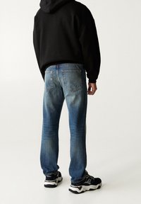 Vaqueros de mezclilla azul claro con un ajuste relajado, que presentan áreas desgastadas, dos bolsillos traseros y detalles con costuras. Combinados con una sudadera negra con capucha y zapatillas.