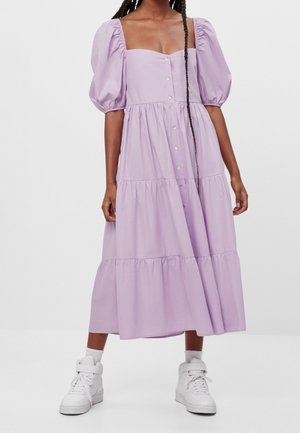 Woman wearing a lilac tiered midi dress with puff sleeves, button front, paired with white high-top sneakers and ankle socks.