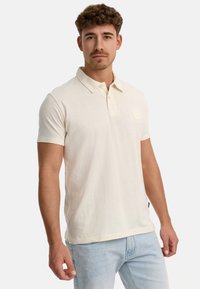 Cremfarbener Poloshirt aus strukturiertem Material, mit kurzen Ärmeln und knöpfbarem Kragen. Mit einem Logo auf der linken Brust. Getragen mit heller Denim.
