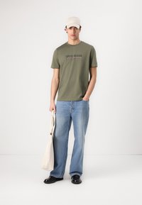 T-shirt de algodão verde com estampa de logotipo, combinada com jeans largos de cor azul clara, sapatos pretos e um boné creme, segurando uma bolsa de lona.