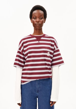 Persoon draagt een bordeauxrood-wit gestreept T-shirt met korte mouwen over een wit topje met lange mouwen en een blauwe spijkerbroek, staand tegen een witte achtergrond.