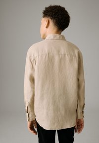 Beige langärmeliges Hemd aus strukturiertem Stoff, mit Kragen und geknöpften Manschetten, getragen mit schwarzer Hose, Rückansicht.