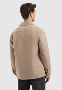 Beige wollen jas met een kraag, rechte snit en knoopmanchetten. Heeft een gestructureerde stof en zichtbare stiksels op de rug.