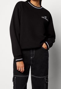 Czarny świetlik z białym prążkowanym dekoltem i mankietami, z białym logo. Zestawiony z ciemnymi spodniami cargo, podkreślającymi luźny krój.