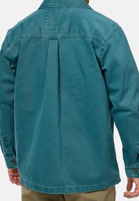 Giacca di denim verde acqua con colletto, piega singola sul retro e polsini con bottoni. Presenta una toppa rettangolare sopra la spalla. Texture liscia.