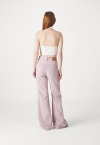 Femme aux longs cheveux roux portant un haut blanc sans bretelles et un pantalon rose taille haute à larges jambes, debout de dos sur fond blanc.