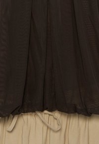 Μαύρο ύφασμα από δίχτυ απλωμένο πάνω σε μπεζ πλισέ υλικό με ορατές κορδόνια δεσίματος στη ραφή μεταξύ των δύο υφασμάτων.