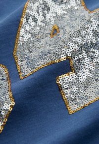 Tela azul con lentejuelas plateadas en forma de estrella y un borde dorado. La textura es suave, con lentejuelas reflectantes y un fondo mate.