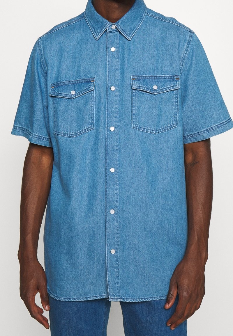 Džínová košile světle modré barvy, krátké rukávy, klasický límec, dvě náprsní kapsy a zapínání na knoflíky vpředu se stříbrnými akcenty. Hladká textura.