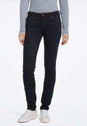 Person iført tætsiddende sorte jeans, stribet langærmet skjorte og hvide sneakers, stående med hænderne afslappet ned langs siderne.