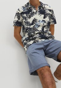 Polo a maniche corte color crema con motivi floreali e fogliari blu, abbinato a pantaloni corti leggeri azzurri arrotolati. Vestibilità casual e tessuto leggero.
