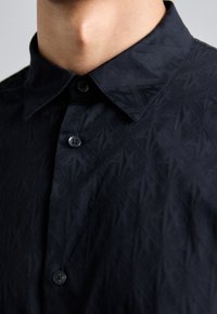 Svart skjorte med knapper som har et teksturert diamantmønster, klassisk krage og matchende knapper, som fremhever en glatt stofffinish.