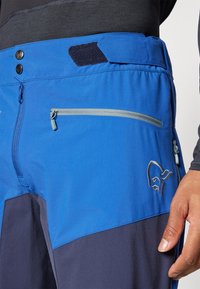 Blue and dark gray outdoor pants featuring a zippered pocket, logo detail, and a velcro waistband, designed for functionality and movement.