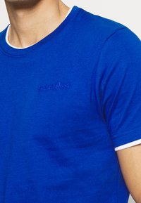 Niebieska bawełniana koszulka z okrągłym dekoltem, kontrastowe białe wykończenie na rękawach oraz haftowane logo po lewej stronie. Gładka faktura.