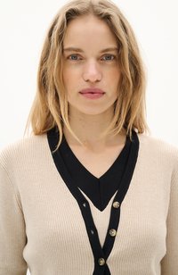 Gilet côtelé beige avec un col en V noir, avec une rangée de trois boutons dorés. Texture lisse, design ajusté et détails subtils.