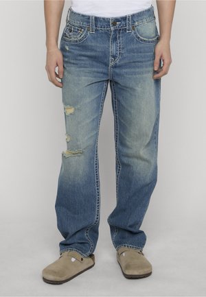 Person trägt hellblaue, abgenutzte Jeans mit sichtbaren Rissen am linken Bein und beige Slip-On-Schuhe vor einfachem Hintergrund.