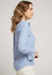 Chemise à rayures bleu et blanche, avec des manches retroussées, fabriquée dans un tissu doux, présentant une coupe décontractée et un ourlet arrondi.