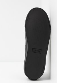 Musta kumikenkien pohja, jossa on tekstuuri kuvio, keskellä logo. Pyöreä muoto ja sileät reunat, minimalistinen muotoilu.