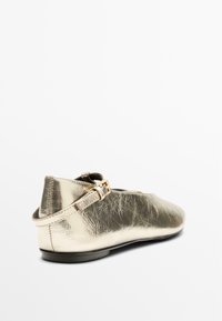 Goldfarbener Ballerinaschuh mit metallischem Finish und strukturierter Oberfläche, ausgestattet mit einem Rückriemen und einer Schnallendekoration. Abgerundete Zehenpartie und flache Sohle.