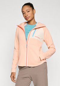 Ελαφρύ μπουφάν σε ροδακινί χρώμα με κουκούλα, με αντίθετο μπεζ πάνελ και λεπτομέρειες από μπλε φερμουάρ, κατασκευασμένο από μαλακό, ελαστικό ύφασμα.