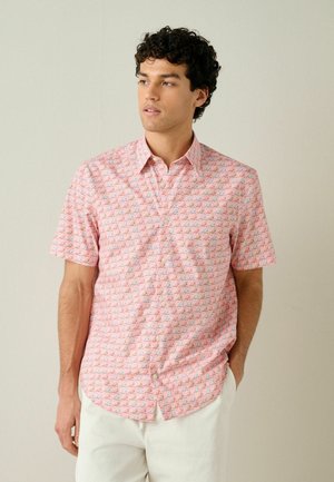 Mann mit lockigem dunklem Haar, der ein hellrosa Hemd mit kurzen Ärmeln und geometrischem Muster sowie eine weiße Hose trägt und mit einer Hand in der Tasche steht.