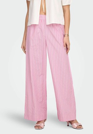 Kobieta ubrana w szerokie spodnie w pionowe różowo-białe paski, białą bluzkę z krótkim rękawem oraz różowe sandały na obcasie z odkrytymi palcami i kokardami.