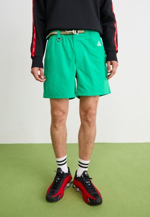 Green shorts with a textured surface, an adjustable gold belt, and a small logo on the side, paired with black and red sneakers and striped socks.