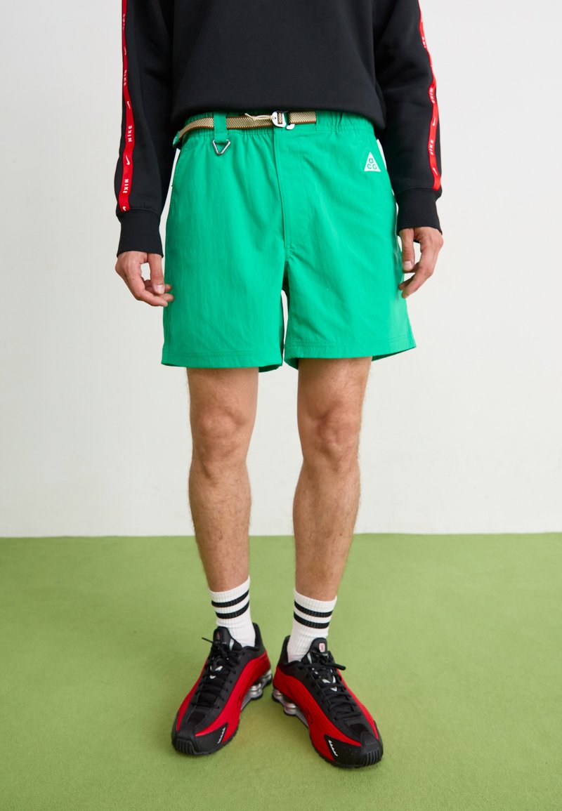 Shorts verzi cu o suprafață texturată, o curea ajustabilă aurie și un mic logo pe lateral, asortați cu teniși negri și roșii și șosete cu dungi.