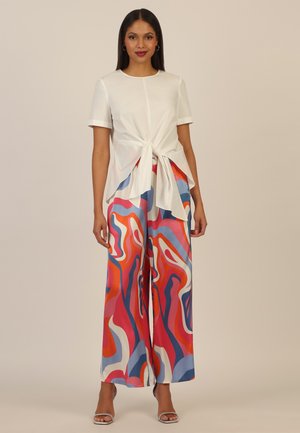 Frau trägt ein weißes kurzärmeliges Oberteil, das an der Taille gebunden ist, kombiniert mit hoch taillierten, weit geschnittenen Hosen mit abstrakten mehrfarbigen Mustern.