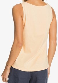 Vrouw in een mouwloze licht perzikkleurige top en donkere marineblauwe broek, van achteren gezien, met een gouden armband om haar linkerpols.