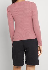 Haut à manches longues rose côtelé avec un col rond, coupe ajustée et texture douce. Associé à un short foncé, laissant apparaître des tatouages visibles sur les jambes.