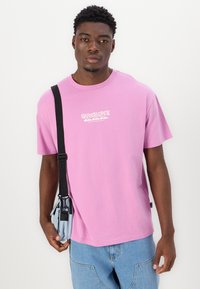 T-shirt en coton rose à manches courtes et col rond ; avec impression d'un logo blanc sur la poitrine. Associé à un pantalon en denim bleu et un petit sac.