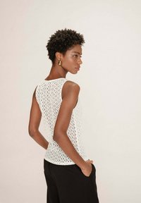 Haut sans manches blanc en tissu texturé avec un motif géométrique ; porté avec un pantalon noir, mettant en valeur le design et l'ajustement au dos.