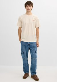 Joven de pie mirando al frente, vistiendo una camiseta beige con la palabra "Yosemite", jeans cargo azules con grandes bolsillos y zapatos marrones con cordones, sobre un fondo blanco.