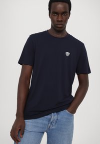 Tamsiai mėlynas medvilninis marškinėliai su apvalia iškirpte, trumpomis rankovėmis ir maža logotipo etikete kairėje krūtinės pusėje. Lygus paviršius ir įprastas prisitaikymas.