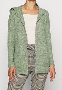 Zelený pletený kardigan s kapucí, dvěma předními kapsami a texturovaným povrchem, v kombinaci s kostkovanými kalhotami a světlým topem.