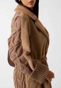 Brązowy, oversize'owy płaszcz o miękkiej, futrzanej strukturze i rękawach z wzorem w warkocze. Posiada kołnierz i szerokie prążkowane mankiety.