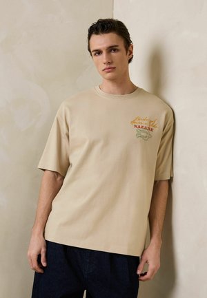 Mladý muž s krátkými tmavými vlasy, který má volné béžové tričko s nápisem "Nazare" a tmavé kalhoty, stojí před texturovanou béžovou zdí.