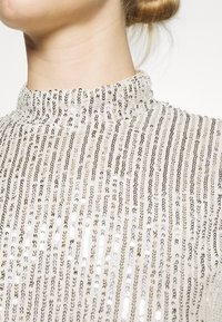 Maglione a collo alto realizzato in un tessuto crema decorato con righe verticali di paillettes argento, creando una superficie testurizzata e riflettente.