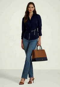 Mulher vestindo uma blusa azul-marinho e jeans azuis, segurando uma bolsa castanha e preta, de pé com as pernas cruzadas e usando saltos castanhos.