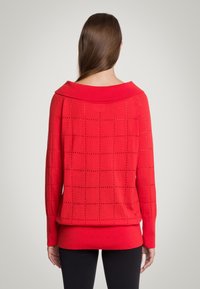 Czerwony sweter dziergany z szerokim kołnierzem, z wzorem w kratę i otwartym ściegiem. Długie rękawy i luźny krój, zestawiony z czarnymi legginsami.