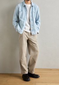 Light blue denim shirt with two chest pockets, white T-shirt underneath, beige pants, and black shoes on wooden floor.