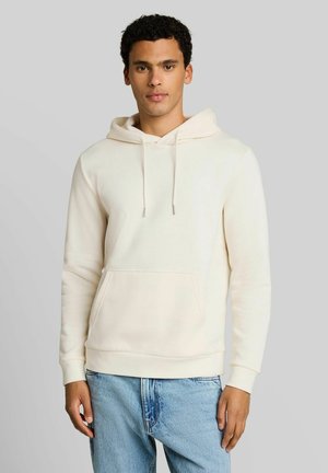 Jonge man draagt een effen off-white hoodie met een voorzak en lichtblauwe spijkerbroek, staand voor een grijze achtergrond.